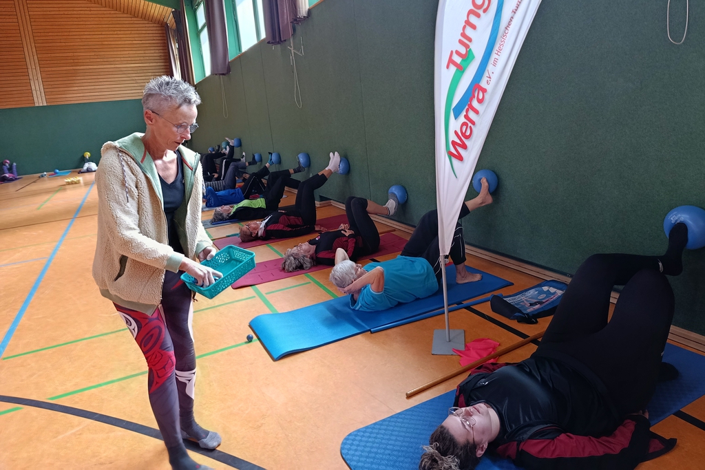Eine Gruppe von Frauen macht gemeinsam eine Übung mit Ball an der Turnhallenwand. 