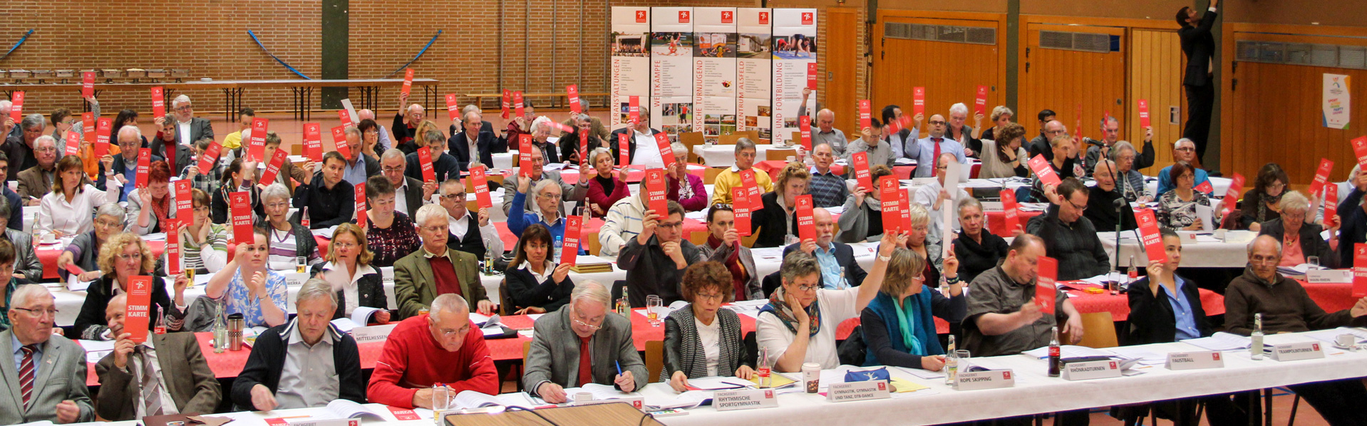 Viele Menschen sitzen an mehreren Reihen von Tischen und halten Stimmkarten nach oben.