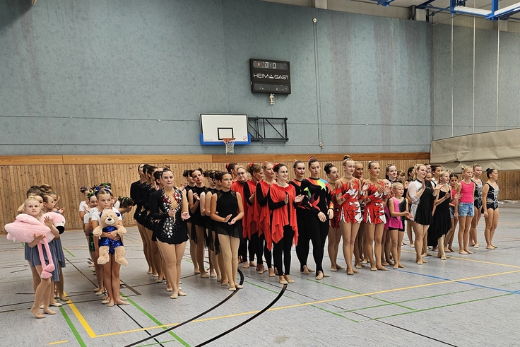 Mehrere Gruppenmitglieder in bunten Kostümen stehend nebeneinander aufgereiht in einer Sporthalle.