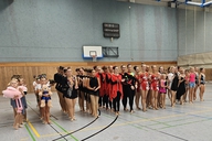 Mehrere Gruppenmitglieder in bunten Kostümen stehend nebeneinander aufgereiht in einer Sporthalle.