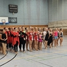 Mehrere Gruppenmitglieder in bunten Kostümen stehend nebeneinander aufgereiht in einer Sporthalle.