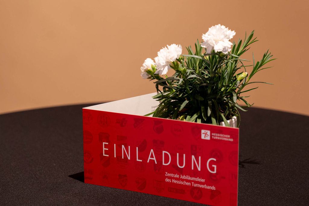 Die Einladungskarte steht vor einer Blume auf dem Tisch, sodass groß "Einladung" zu lesen ist