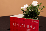 Die Einladungskarte steht vor einer Blume auf dem Tisch, sodass groß "Einladung" zu lesen ist