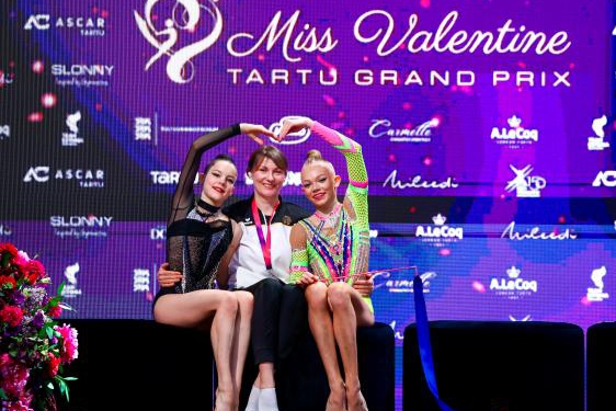 Zwei Gymnastinnen sitzen links und rechts von ihrer Trainerin und bilden mit ihren Armen ein Herz