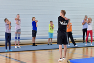 Ein Mann macht mit Kindern eine Dehnübung in der Turnhalle.