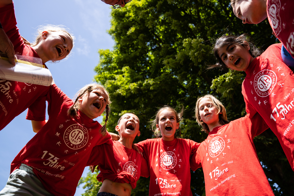 Kinder stehen in einem Kreis, legen die Arme umeinander und rufen einen gemeinsamen Spruch.