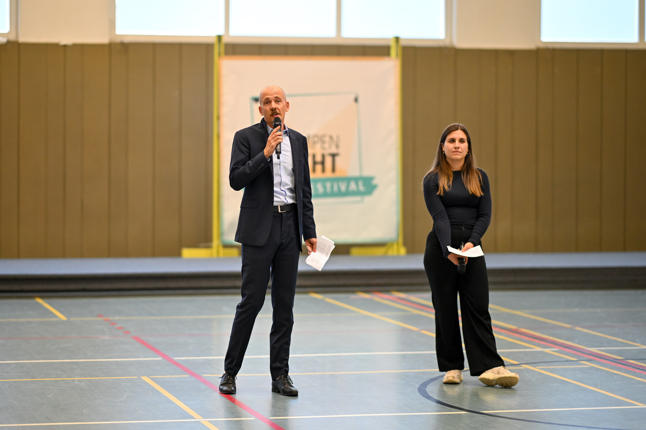 Ein Mann spricht in ein Mikro, daneben steht eine junge Frau schaut zu.