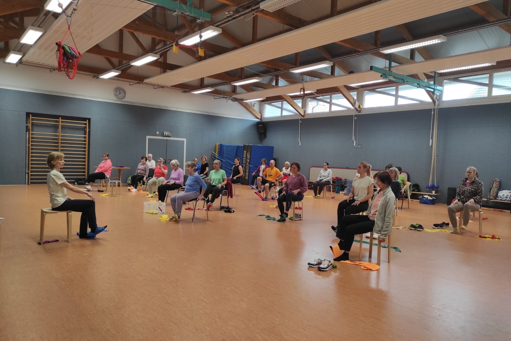 Eine Gruppe sitzt in einer Sporthalle und zeigt eine Sportübung.