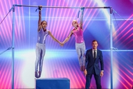 Zwei Turnerinnen hängen einarmig am Reck, davor steht der Moderator Kai Pflaume