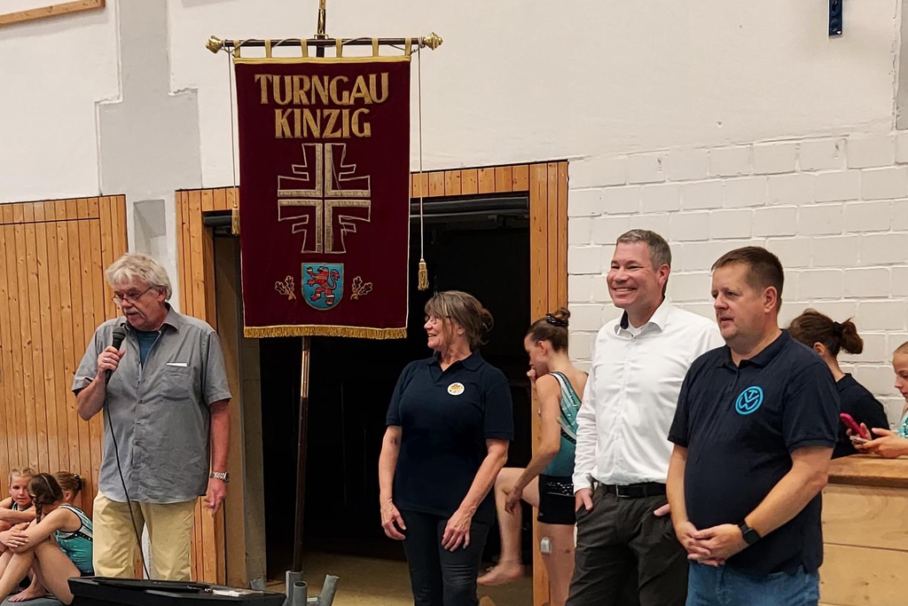Vier Personen stehen vor einem Banner mit der Aufschrift "Turngau Kinzig", eine Person mit Mikro.