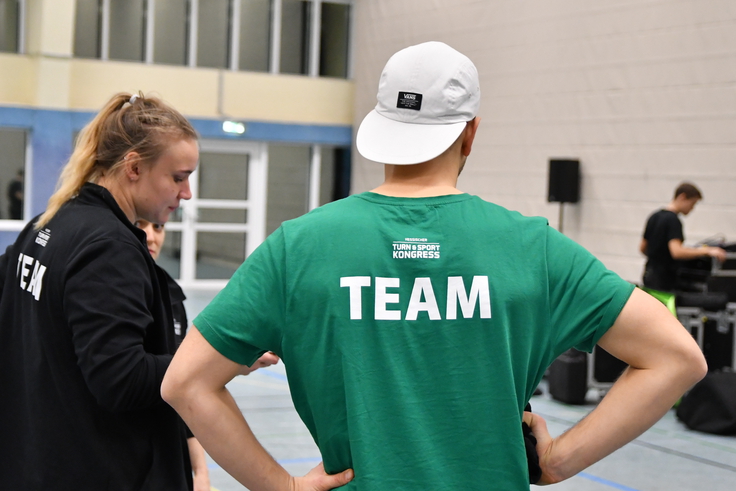 Ein Mann steht in einer Sportgalle und träft die Rückenaufschrift "TEAM".