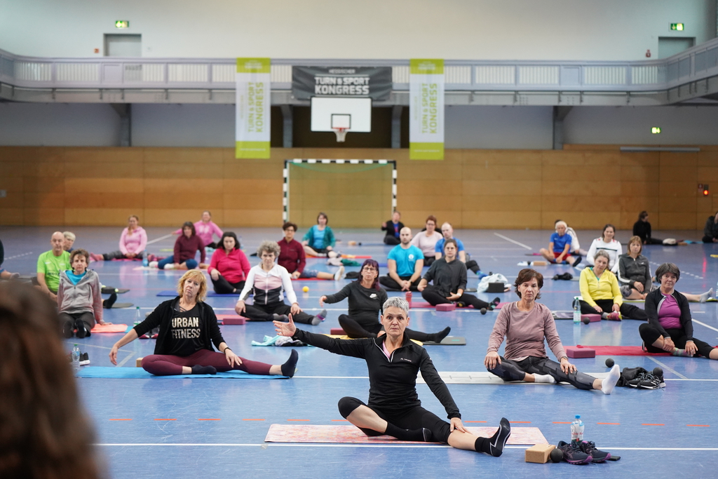 Teilnehmende eines Fitnesskurses sitzen auf ihren Gymnastikmatten und machen eine Übung.