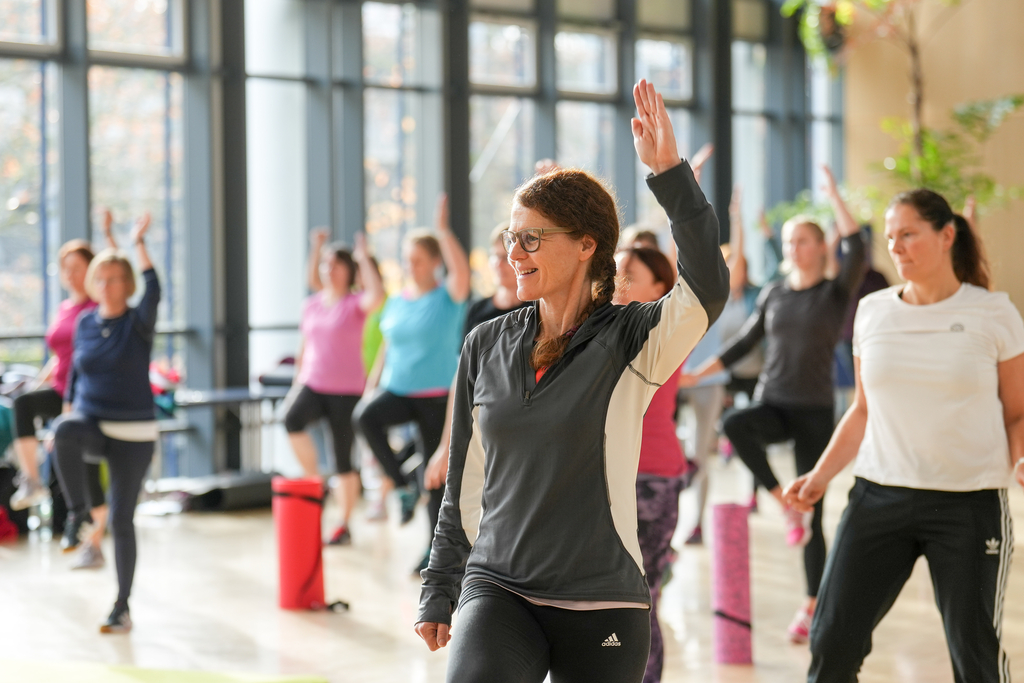 Eine Frau in Sportkleidung hebt die Hand, im Hintergrund sind weitere Personen.
