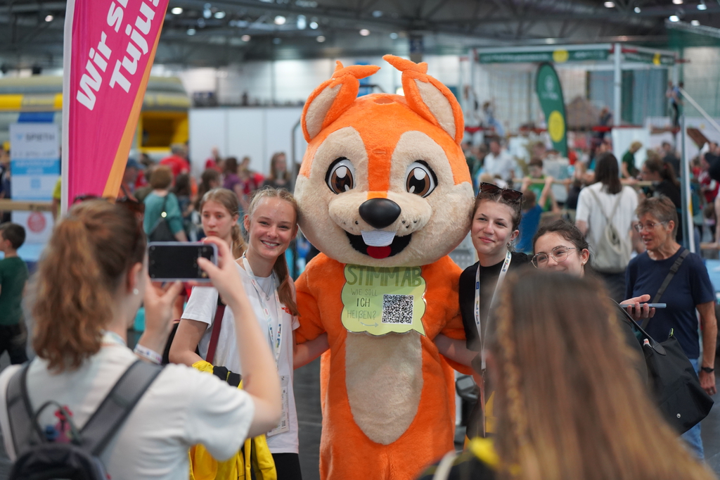 Das neue Maskottchen der HTJ posiert mit zwei jungen Frauen für ein Foto.