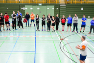 Sportler und Sportlerinnen der Sportart Zweier-Prellball lauschen einem Referierenden in einer Sporthalle.