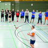 Sportler und Sportlerinnen der Sportart Zweier-Prellball lauschen einem Referierenden in einer Sporthalle.