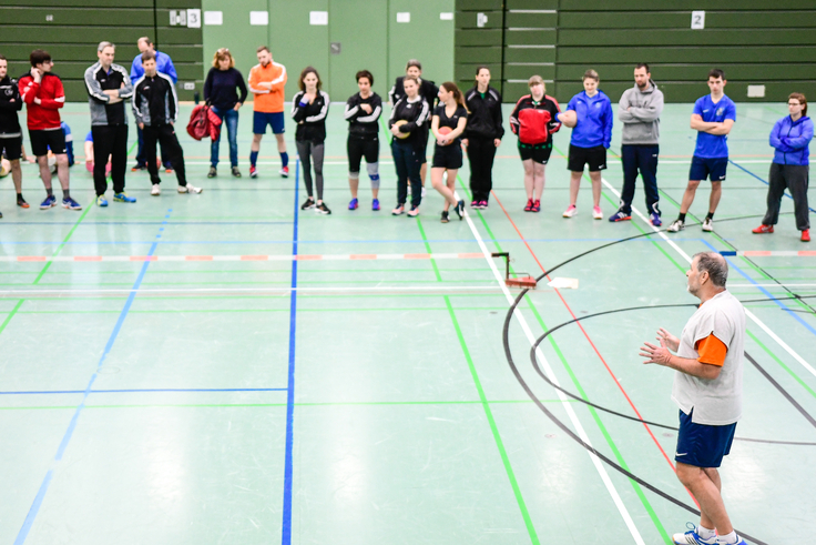 Sportler und Sportlerinnen der Sportart Zweier-Prellball lauschen einem Referierenden in einer Sporthalle.