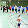 Sportler und Sportlerinnen der Sportart Zweier-Prellball lauschen einem Referierenden in einer Sporthalle.