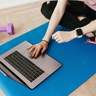 Person sitzt auf einer Sportmatte mit Laptop, daneben liegen Hanteln