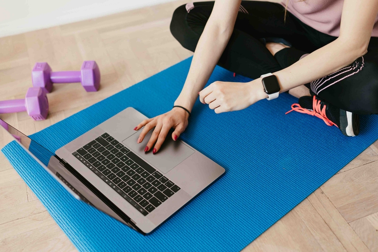 Person sitzt auf einer Sportmatte mit Laptop, daneben liegen Hanteln