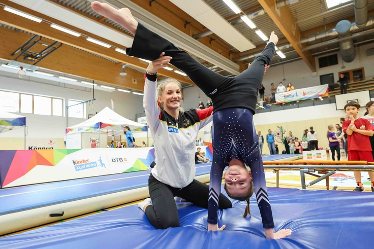 Eine Athletin in der Hocke hält ein Kind beim Handstand.