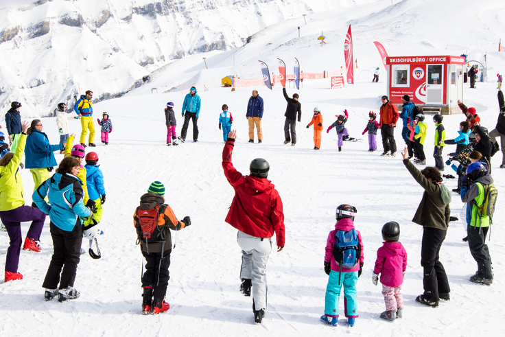 Eine große Gruppe von Skifahrern stehen auf der Schneepiste in einem Kreis und machen gemeinsam Gymnastikübungen.