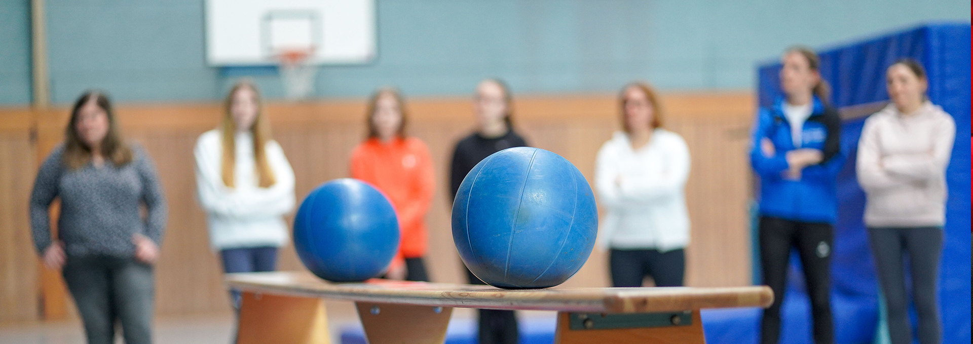 Eine Turnbank mit zwei Bällen darauf, im Hintergrund steht eine Gruppe von Personen.