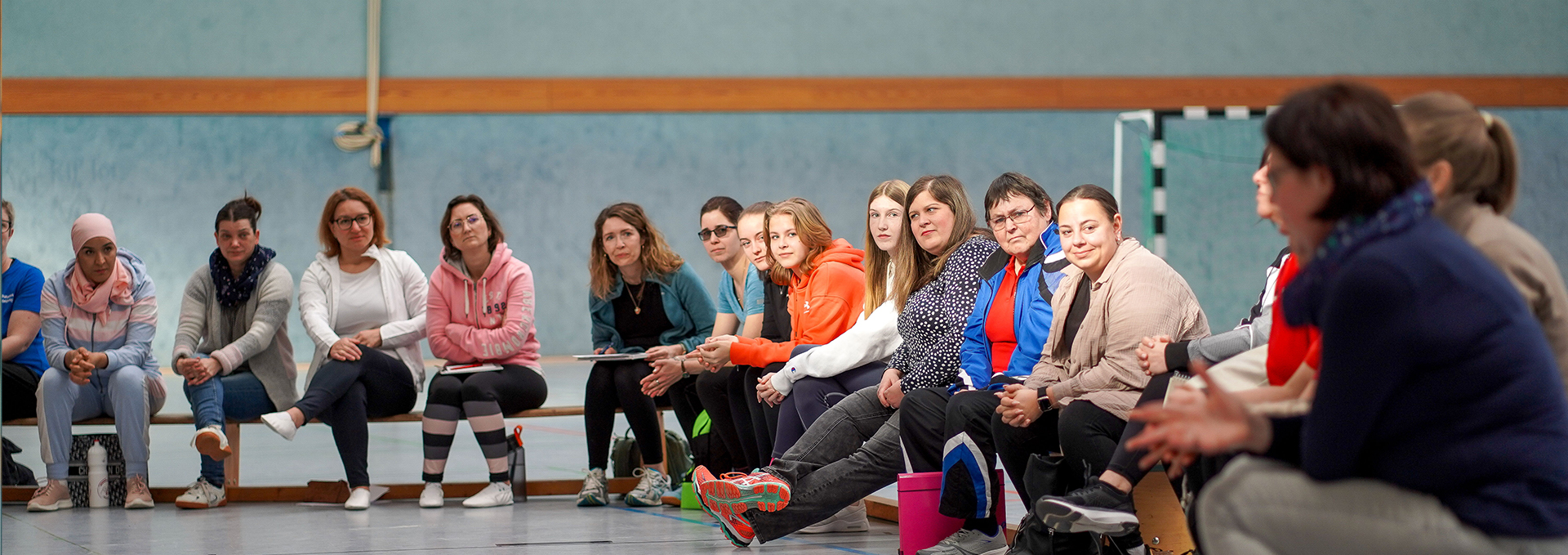 Eine Gruppe sitzt auf Turnbänken und hört einer Person zu.