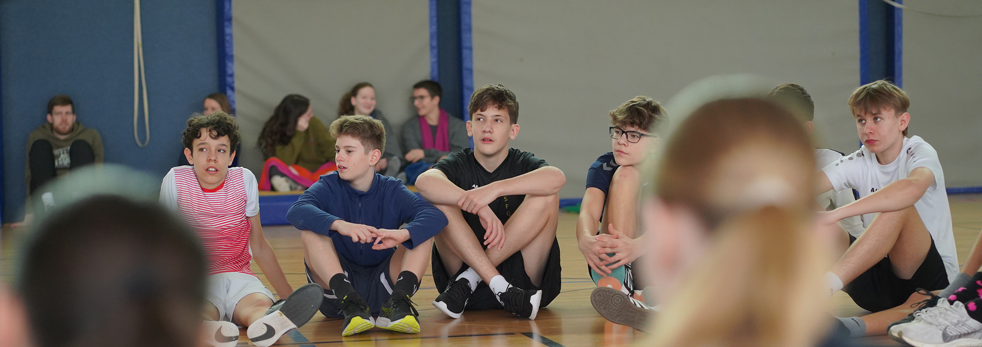 Eine Gruppe schon Schülerinnen und Schüler sitzen in einer Turnhalle im Kreis.