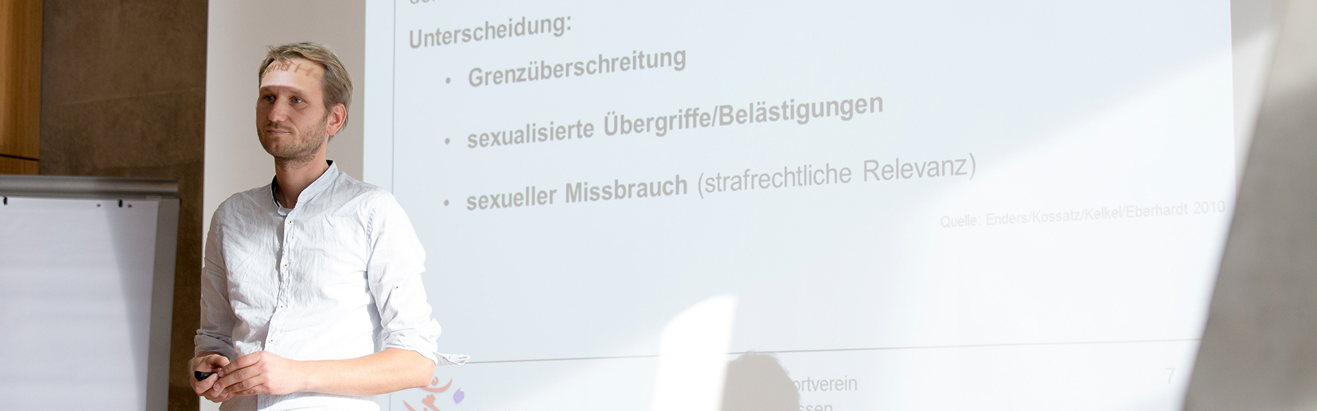 Ein Mann steht vor einer weißen Wand auf der eine Präsentation geworfen wurde.