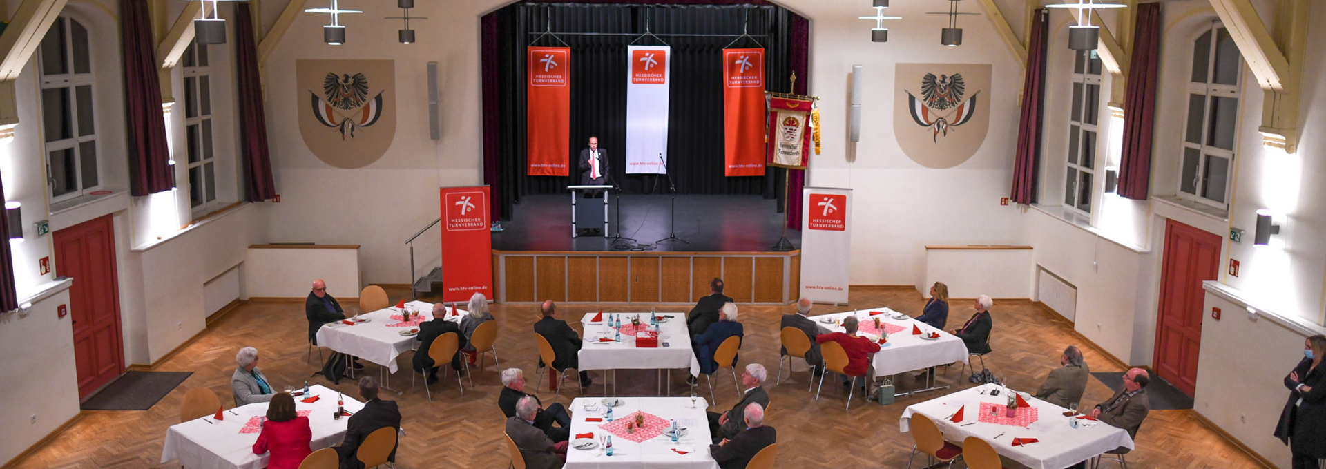 In einem großen Saal sitzen mehrere Personen an Gruppentischen, auf der Bühne hält jemand eine Rede.